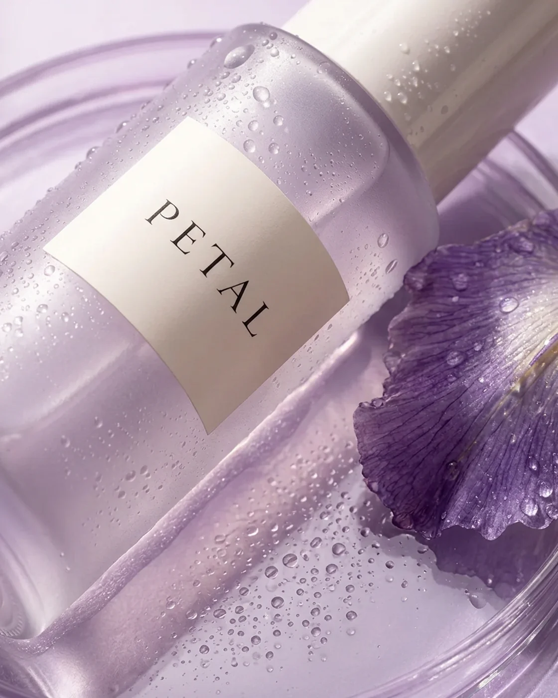 A close diagonal view of the Petal label and frosted lavender glass covered in water beads, beside a purple iris petal.